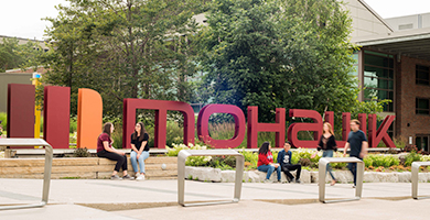Students sitting and walking by the sign at the entrance to Mohawk College's Fennell Campus