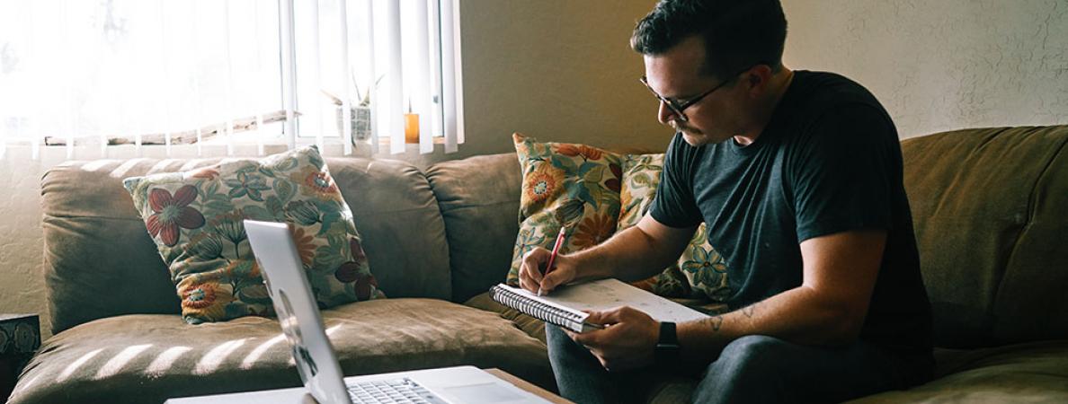 white male taking notes while sitting on couch with laptop open