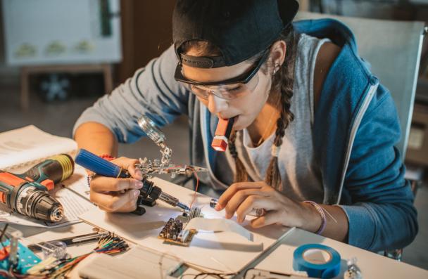 female late teens or early 20s working on soldering metal and wires while sitting at table.
