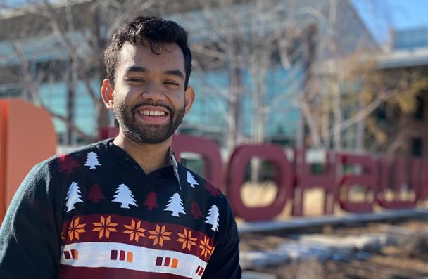 Mirza standing in a holiday sweater in front of the Mohawk College sign
