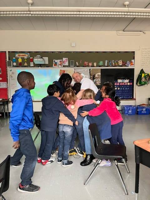 Jay in a classroom surrounded by 8 students giving him a hug. 