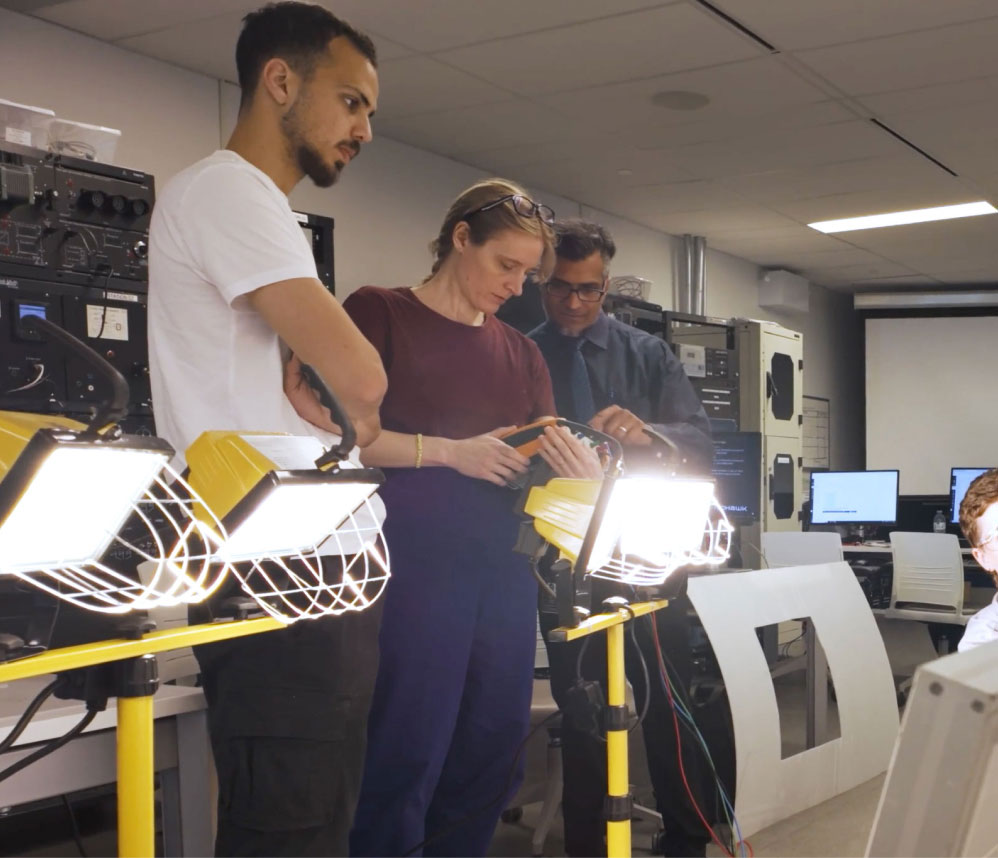 Two Mohawk College students and a faculty member inspecting a portable floodlight in a classroom