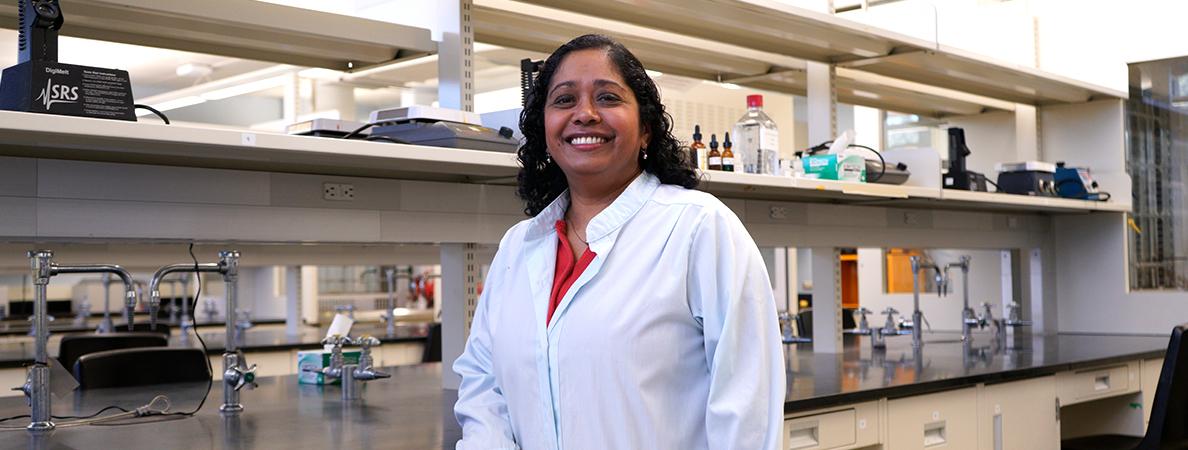 Dr. Vivegananthan smiles as she stands in front of a lab bench filled with professional equipment.