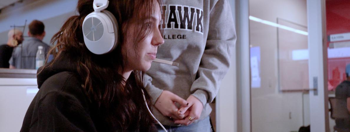 A seated Mohawk College student with long red hair looks at a computer screen outside of the picture. They are wearing a white wired headset. Behind them stands a classmate wearing a grey sweater. The sweater reads 'Mohawk College'.