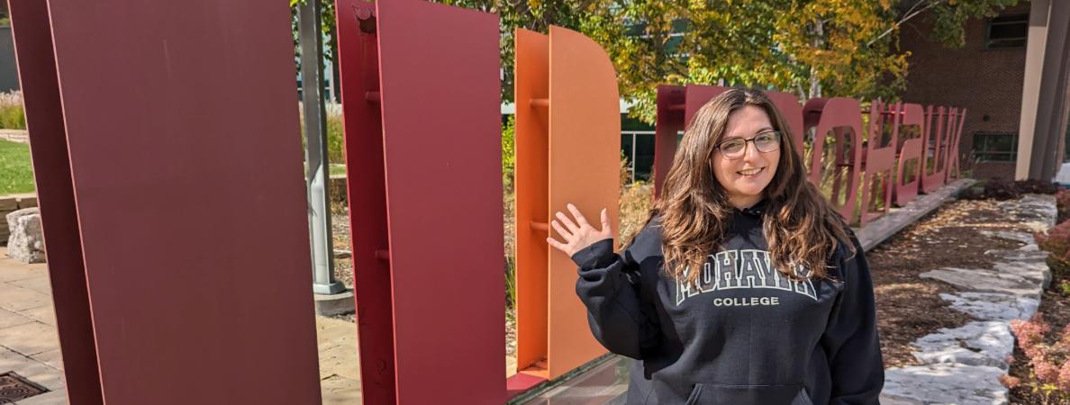 Maggie stands and waves in front of the Mohawk College logo found at the Fennell Campus.