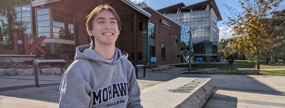 Jonah smiles as he sits in front of the Mohawk College logo at Fennell Campus.
