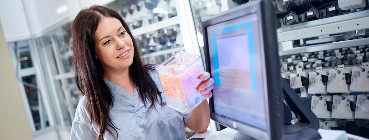 female pharmacy technician reviewing container of drugs standing beside computer