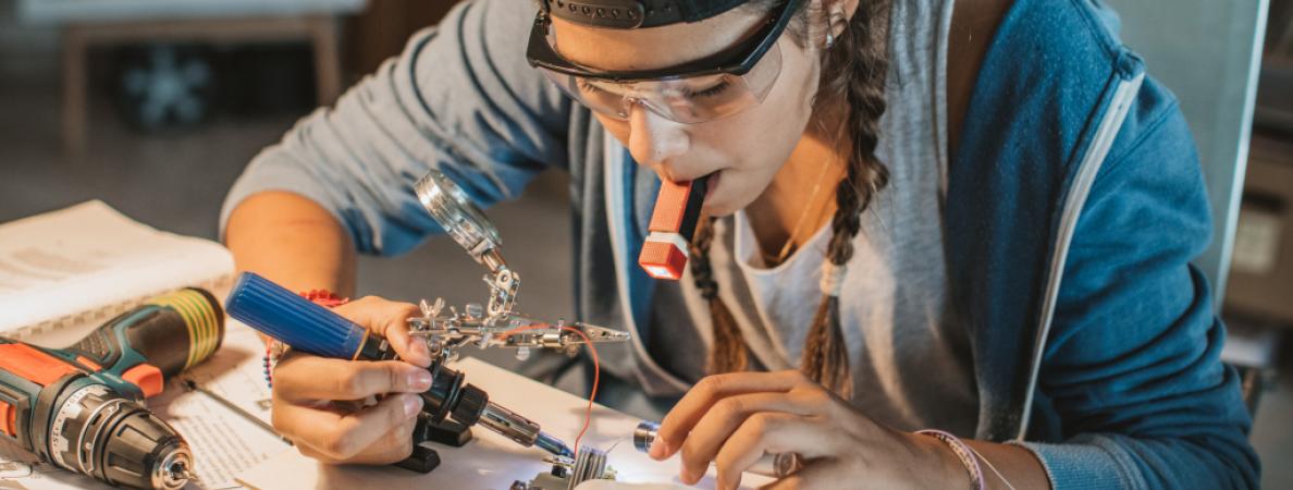 female late teens or early 20s working on soldering metal and wires while sitting at table.