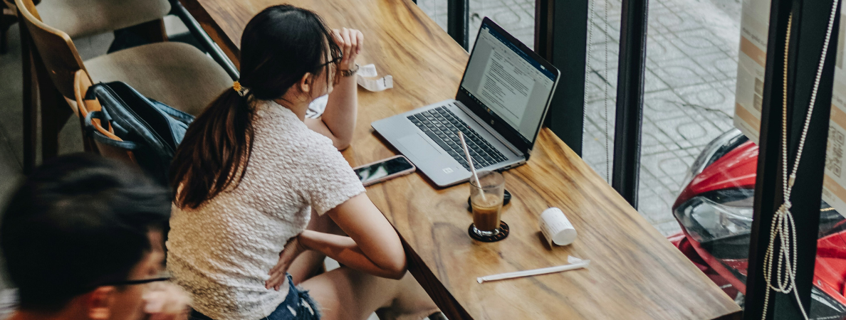 An image of a woman sitting at a table in a coffee shop, looking intently at her laptop.