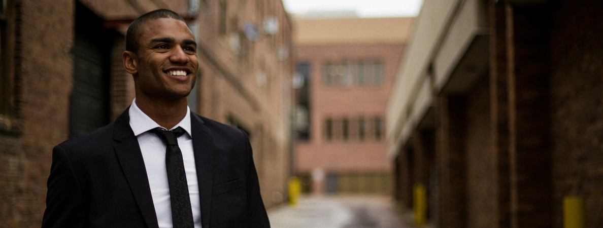 A darker skinned man in a suit with a smile on his face, standing in an urban area