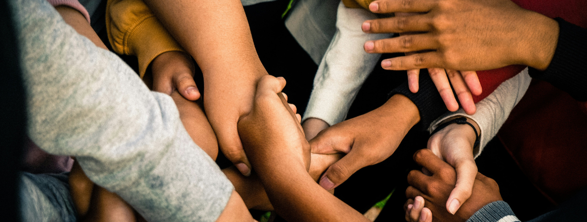 An image of a group of hands, all reach across each other to hold another hand.  The hands are different sizes and skin tones.