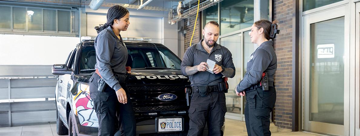 Three Mohawk Advanced Police Studies students in front of a police car talking.