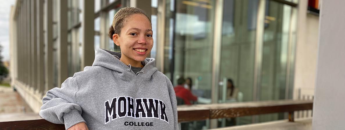 Korina in a Mohawk College hoodie standing in front of the library's exterior