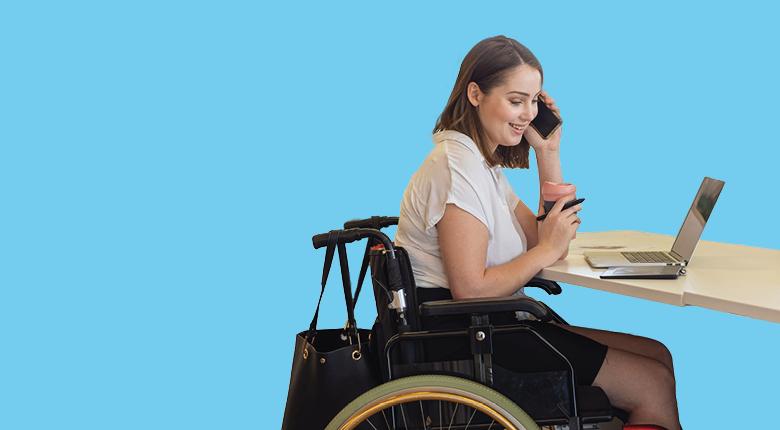 A woman in a wheelchair is speaking on the phone and using her laptop on the table.