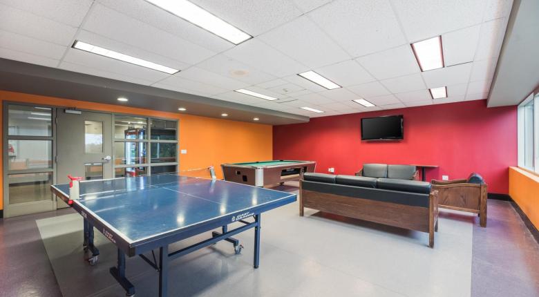 View of the games lounge with bright orange and red walls, a ping pong table, pool table and couches in front of a flat screen TV.