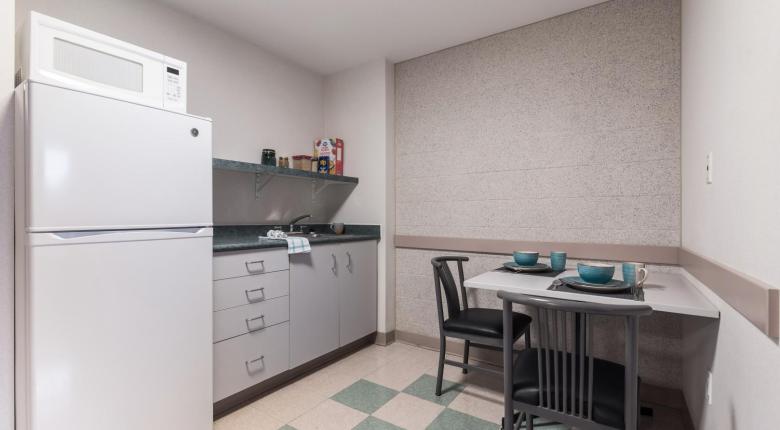 Suite kitchenette with a microwave on top of a full sized fridge, a countertop with sink, and a table with 2 chairs.
