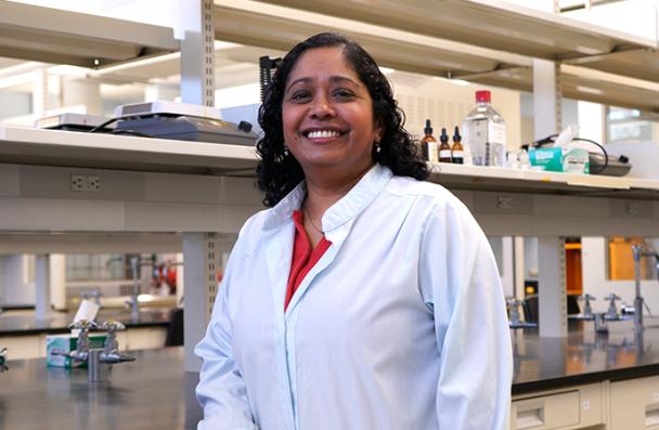 Dr. Vivegananthan smiles as she stands in front of a lab bench filled with professional equipment.