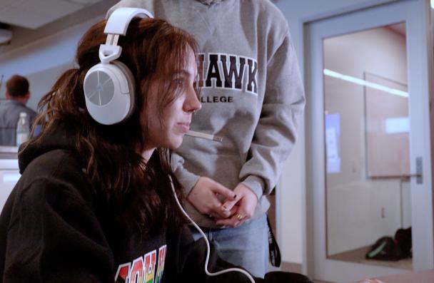 A seated Mohawk College student with long red hair looks at a computer screen outside of the picture. They are wearing a white wired headset. Behind them stands a classmate wearing a grey sweater. The sweater reads 'Mohawk College'.