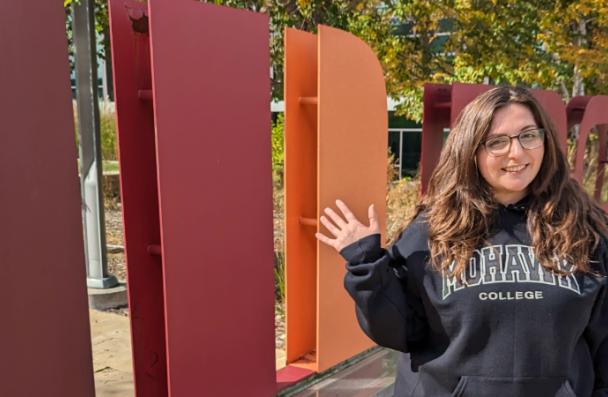 Maggie stands and waves in front of the Mohawk College logo found at the Fennell Campus.