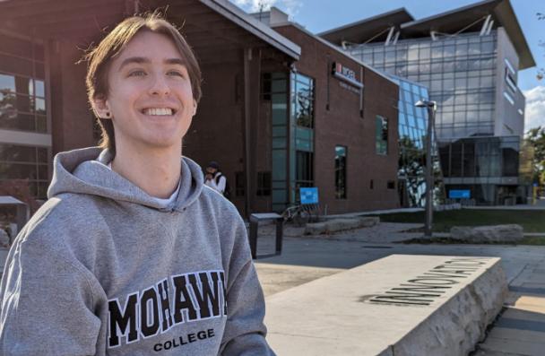 Jonah smiles as he sits in front of the Mohawk College logo at Fennell Campus.