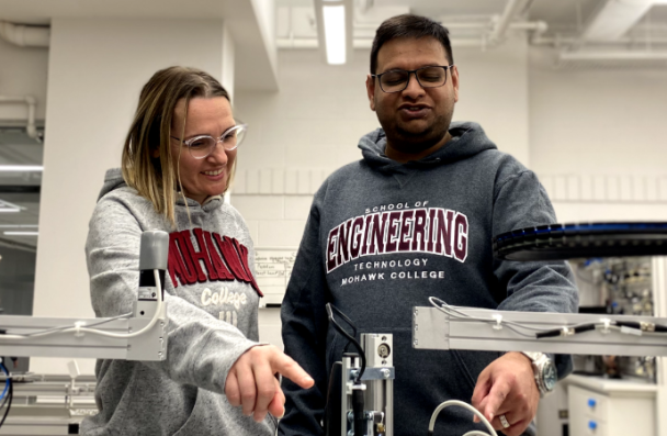 Two Mohawk College students stand amongst a wide array of robotics equipment.