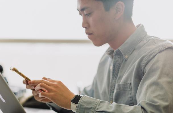 asian male lookig at phone with laptop open while sitting at table. 